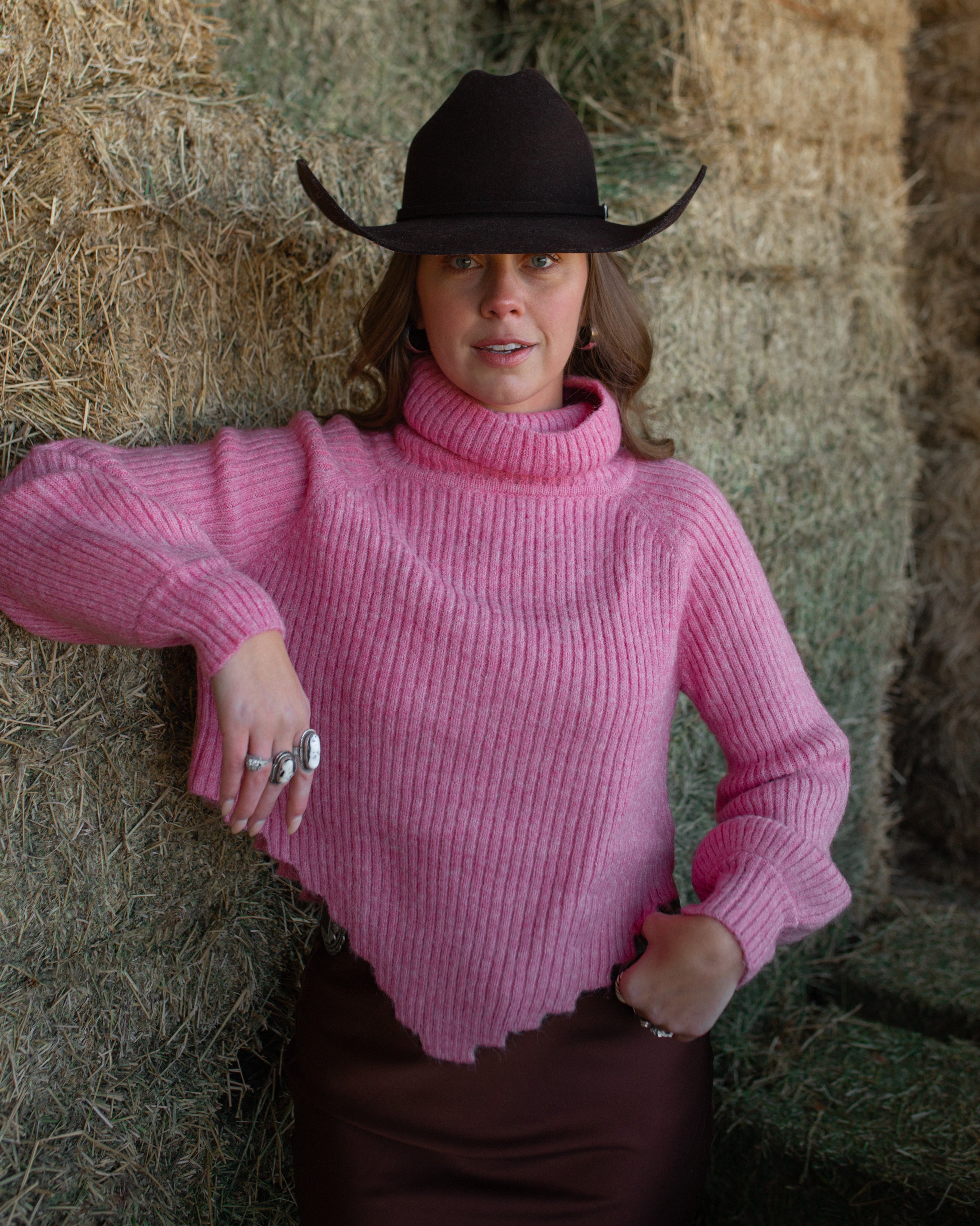 Woman wearing a pink sweater and black cowboy hat standing in front of hay bales.